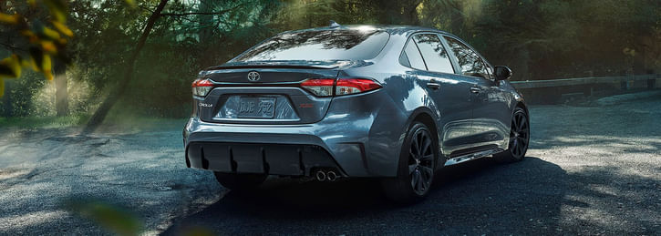 A sleek silver 2026 Toyota Corolla parked in a forested area, showcasing its sporty rear design and dual exhausts. Sunlight filters through the trees.