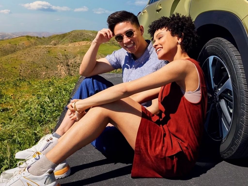 A man and woman sitting together on the side of a parked car, smiling and enjoying each other's company.