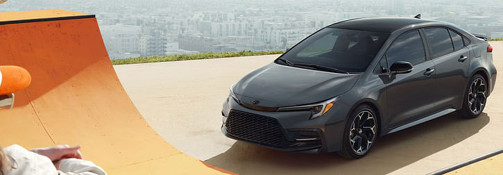 A gray 2025 Toyota Corolla parked next to a bright orange skate ramp, with a cityscape visible in the background.