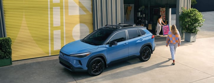 Blue compact SUV parked outside a garage with a large yellow door, while two people walk nearby, one carrying a framed artwork.