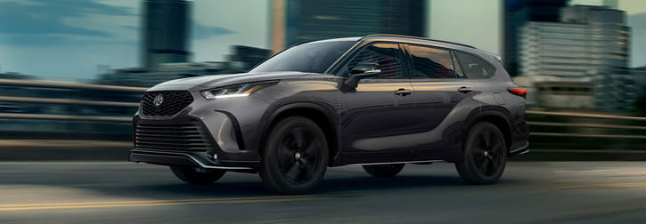 Front-side view of a sleek 2026 Toyota Highlander Hybrid SUV cruising through an urban city landscape at dusk.