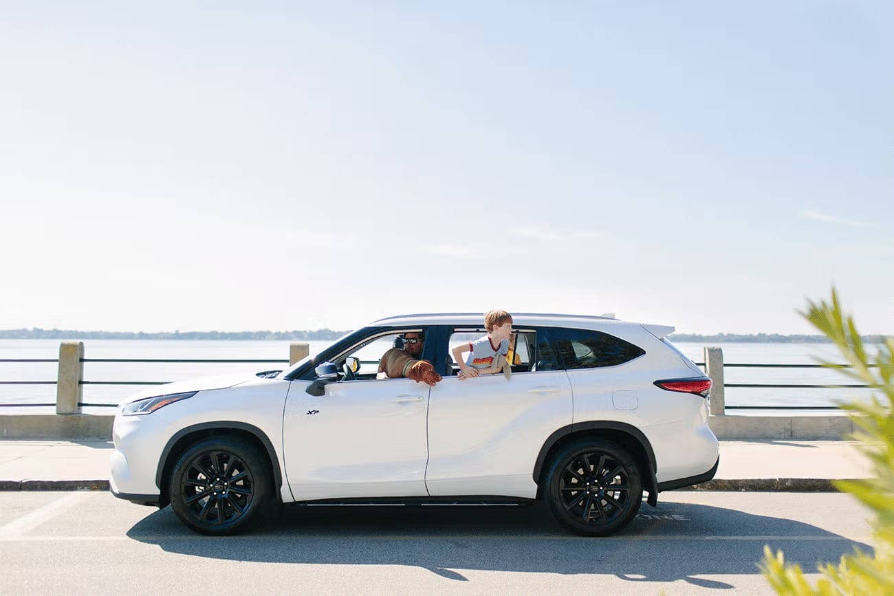 Boy and man leaning out of windows of a parked white SUV by waterfront on a sunny day.
