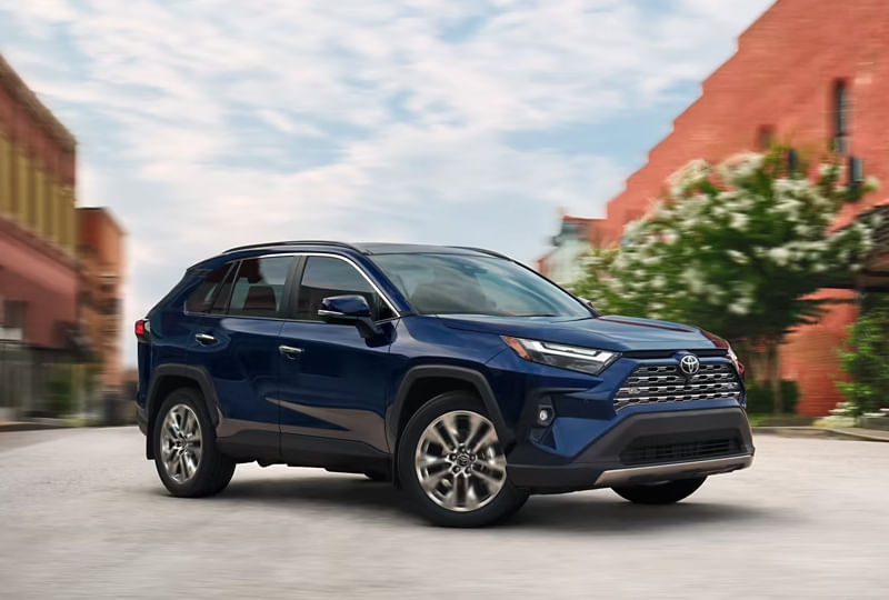 A dark blue Toyota RAV4 SUV driving on a city street, with blurred background indicating motion and red brick buildings on the side.