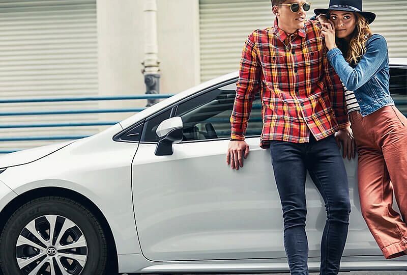 Young couple leaning against white 2022 Corolla Hybrid parked on street in front of sidewalk railing