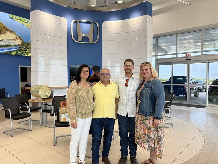 Four people stand smiling in a car dealership showroom. A large Honda logo is prominently displayed on a blue and white wall behind them.
