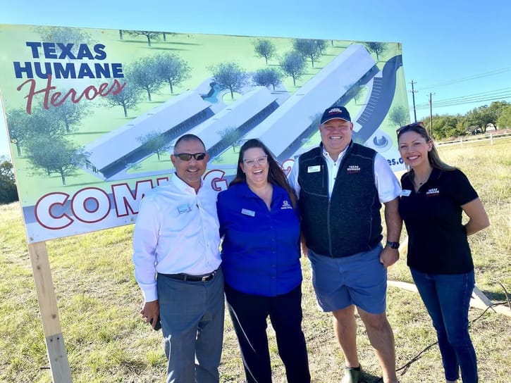Four people smiling in front of a sign that reads "Texas Humane Heroes" with a future building plan. The mood is cheerful and optimistic.