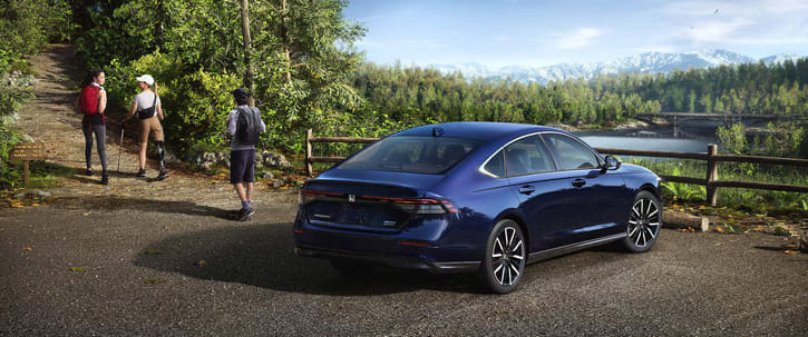 Dark blue 2025 Honda Accord, viewed from the rear passenger side, parked on asphalt near a hiking trail entrance, with a scenic background of a lake, forested hills, and mountains.