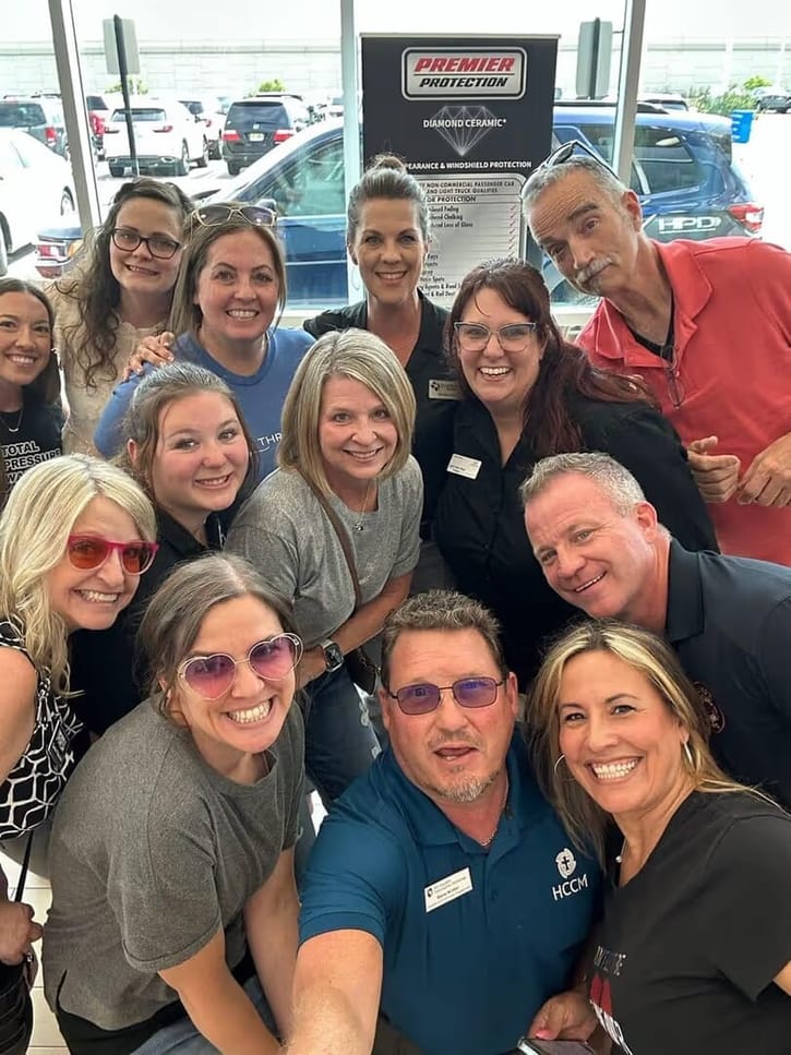 Group of people smiling and posing for a selfie indoors near a car dealership display with vehicles visible outside.