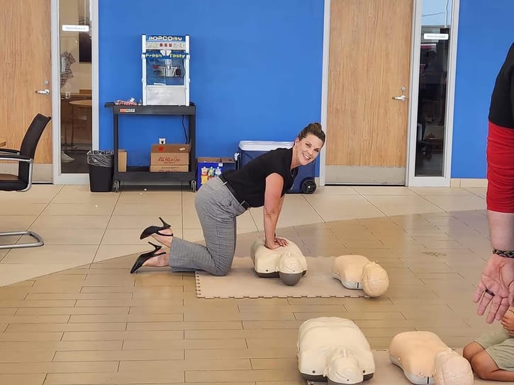 A woman kneels, performing CPR on a dummy in a bright room with a blue wall. She appears focused and calm, with additional dummies nearby.