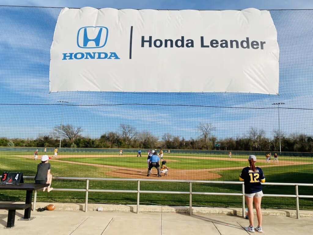 Baseball game under a 'Honda Leander' banner on a sunny day. A player in a blue jersey stands by the railing, with the game in action on the field. Scoreboard shows 3-4.