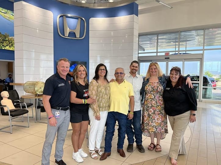 A group of seven people stands smiling in a Honda dealership, with the brand logo on a blue wall behind them. The mood is friendly and welcoming.