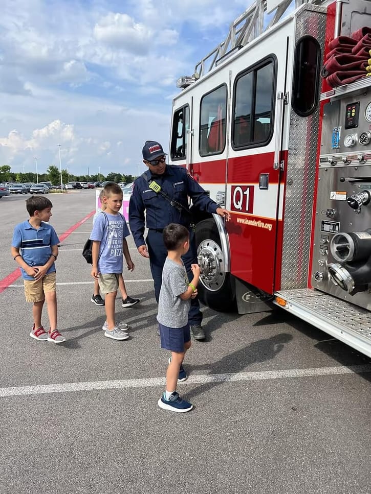 A firefighter in uniform shows a group of young boys around a red fire truck in a parking lot. The children appear curious and engaged.