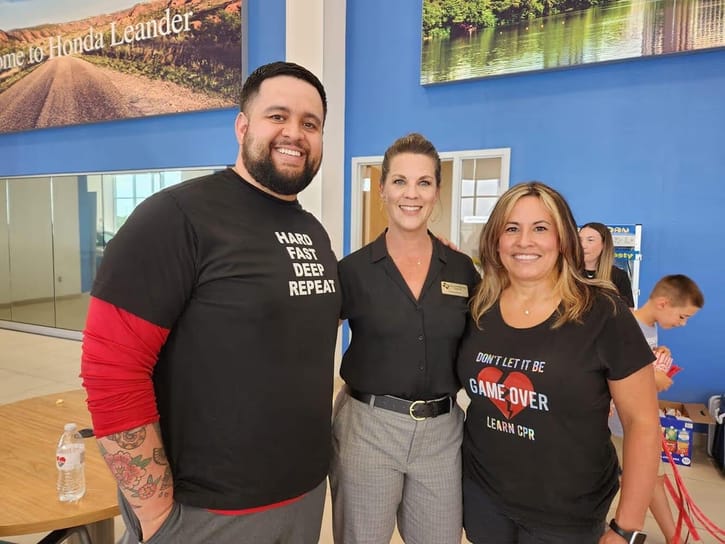 Three people standing indoors near a table, two wearing black t-shirts with motivational CPR messages, with a blue wall in the background.