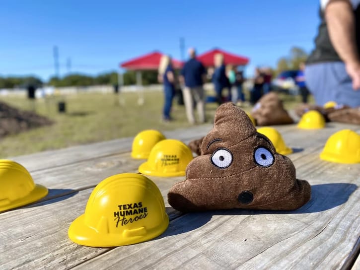 Soft plush poop emoji with cartoon eyes sits among small yellow helmets labeled "Texas Humane Heroes" on a wooden table outdoors; people and tents in the background.