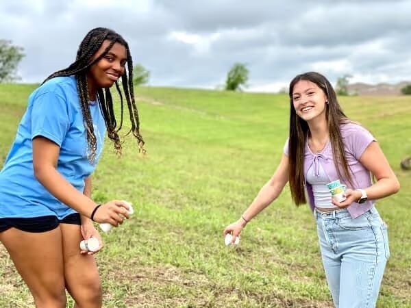 Two young women outdoors on grassy hill, one holding a small cup and both tossing what appears to be marshmallows or similar items.