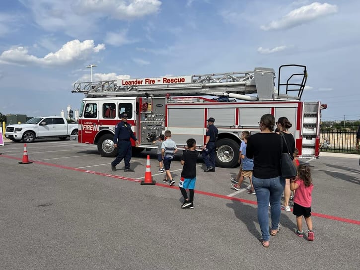 Children and adults explore a red fire truck labeled "Leander Fire - Rescue" in a sunny parking lot. Firefighters engage with the curious crowd.