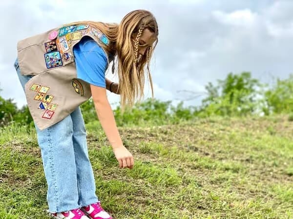 Girl wearing a scout vest with patches, blue shirt, and jeans, bending down to inspect grass in an outdoor grassy area under a cloudy sky.