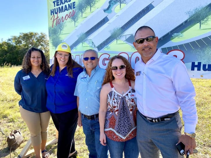 Five people stand smiling in front of a "Texas Humane Heroes" sign on a sunny day. They appear cheerful and united, suggesting a community event or project.