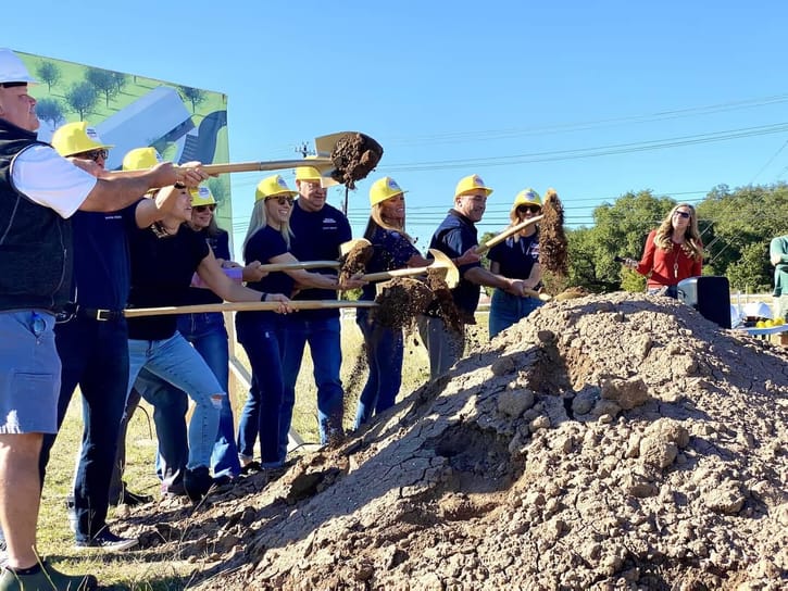 A group of people in yellow hard hats perform a ceremonial groundbreaking, smiling as they lift soil with shovels. The mood is celebratory under a clear blue sky.