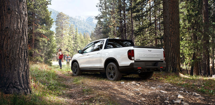 A white 2026 Ridgeline parked on a forest trail as two people set off on a hike among the trees.