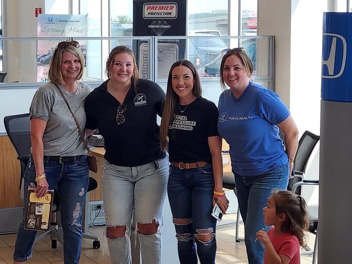 Four smiling women stand together in a Honda dealership. Each wears a branded T-shirt. A young girl looks up at them in the foreground. Bright and cheerful setting.
