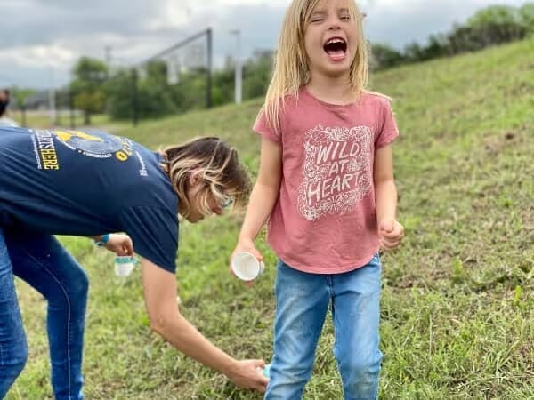 A young girl in a pink "Wild at Heart" t-shirt joyfully laughs on a grassy hill, holding a cup, with an adult near her, reaching down. The scene is playful.