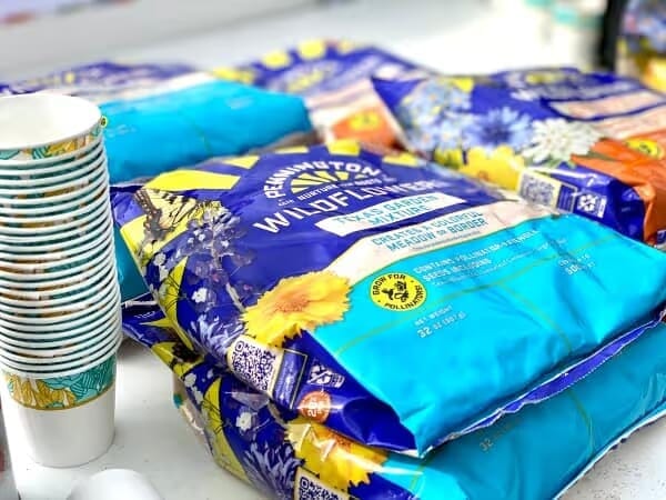 Bags of Pennington wildflower seed mix with colorful floral designs are on a table next to a stack of patterned paper cups. The mood is vibrant and lively.