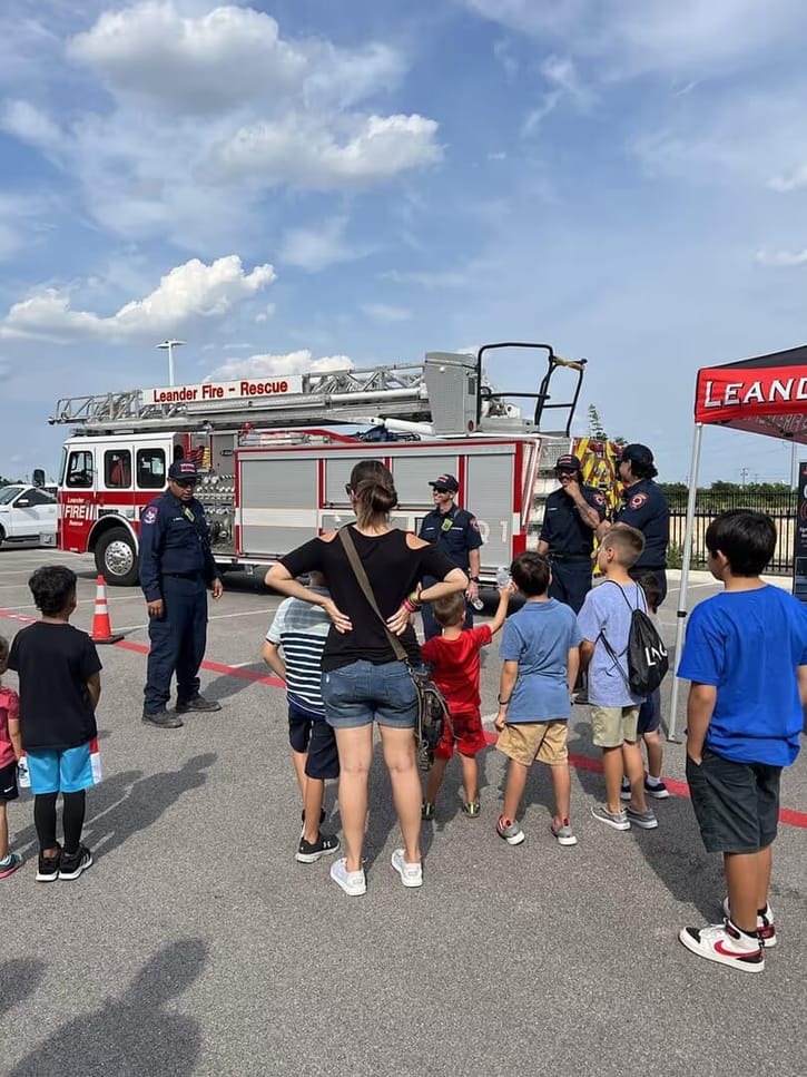 A group of children and an adult stand in front of a fire truck labeled "Leander Fire - Rescue." Firefighters interact with them under a cloudy sky.