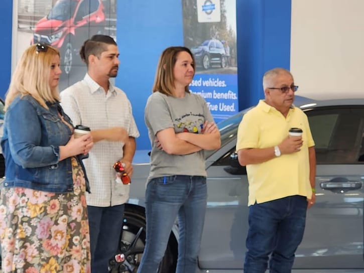 Four people stand in a car showroom, holding coffee cups. They appear attentive and engaged, with a car and promotional posters in the background.