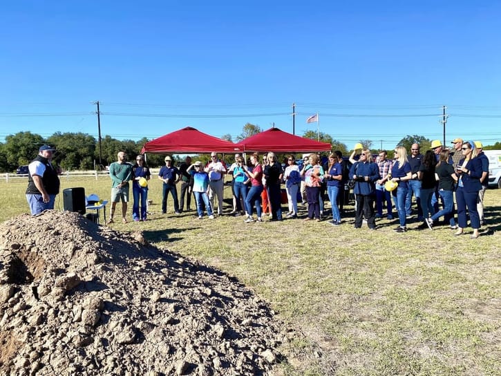 A group of people stands near a dirt pile at an outdoor groundbreaking event, under red tents. A clear blue sky and American flag are visible.