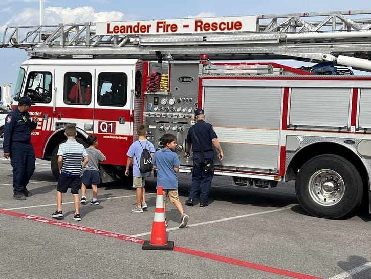 A fire truck labeled "Leander Fire - Rescue" is parked with its side panel visible. Four children are approaching, accompanied by two firefighters. The scene is calm and educational.
