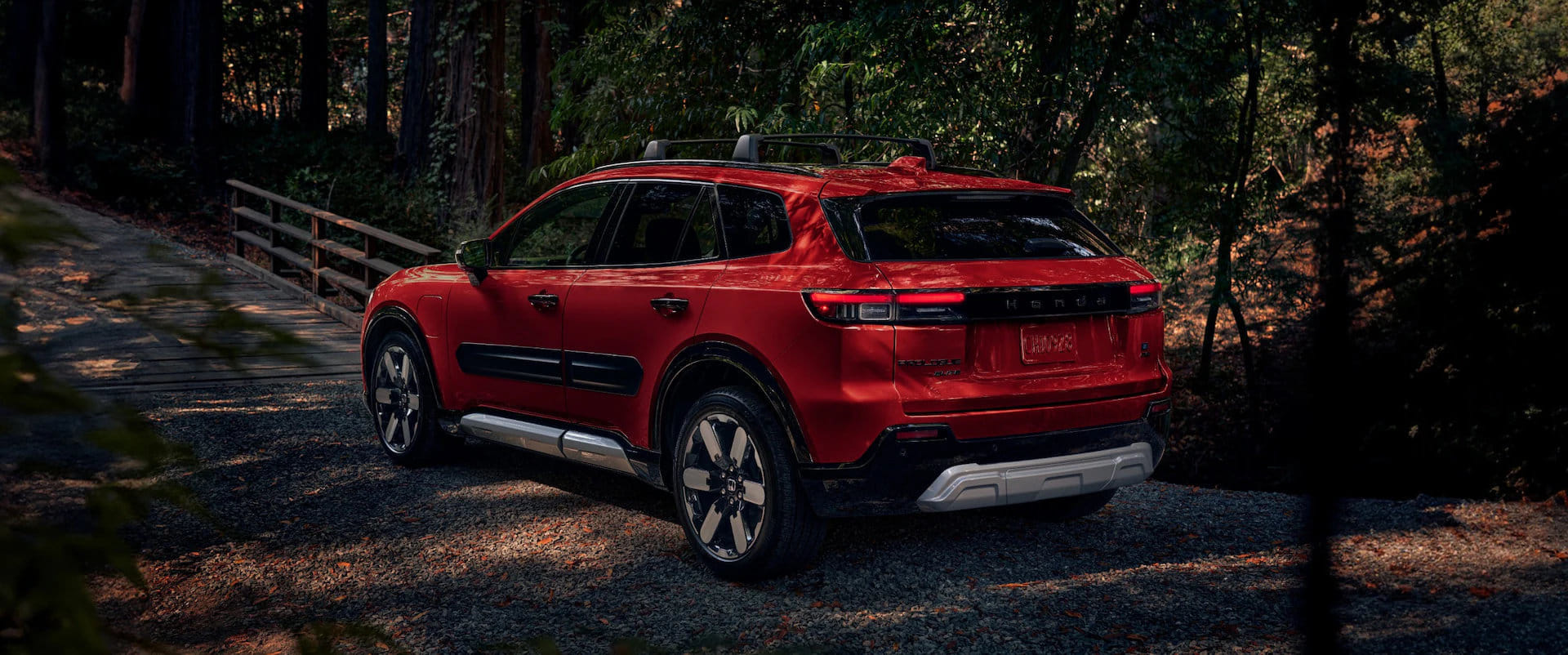 Red 2025 Honda Prologue Elite SUV with a roof rack, parked on a gravel path near a wooden footbridge in a sun-dappled forest