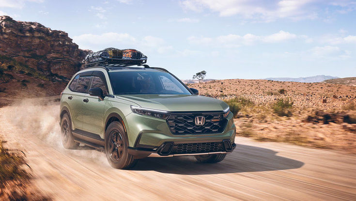 A green Honda CR-V with a roof rack driving on a dusty dirt road in the desert.