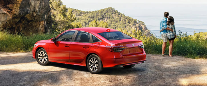 A red Honda Civic sedan is parked on a dirt road at a scenic overlook. To the right, a couple embraces while looking out over a tree-covered hilly landscape towards a large body of water, with a rocky cliff face to the left of the car.