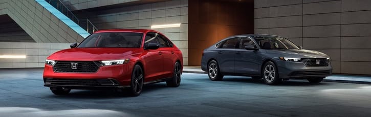 Two 2026 Honda Accord Hybrid are parked indoors on a sleek, modern showroom floor. One car is red, and the other is gray, both featuring stylish, streamlined designs.