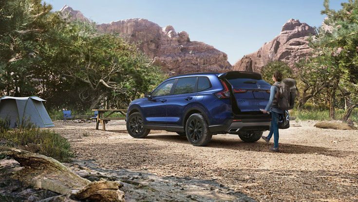 A blue Honda CR-V parked at a campsite with a person unloading gear from the open trunk.