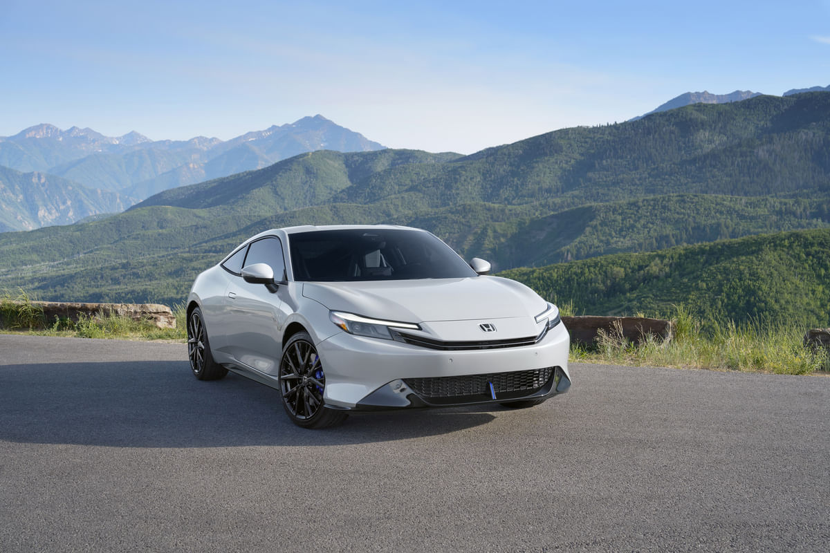 A front three-quarter view of the silver 2026 Honda Prelude parked on a mountain overlook, with a panoramic view of green hills and distant peaks under a blue sky.
