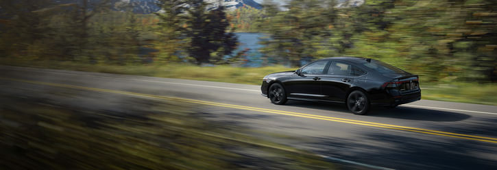 A sleek, black 2025 Honda Accord Hybrid speeds along a scenic road, surrounded by vibrant autumn foliage and a serene lake in the background.