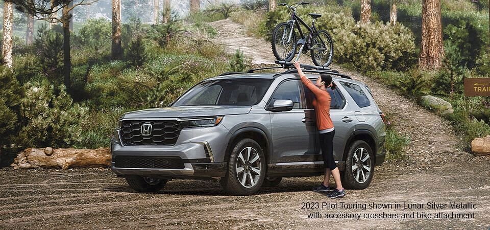 Woman placing bicycle on 2024 Honda Pilot shown in Lunar Silver metallic with accessory crossbars and bike attachment standing on a forest parking.
