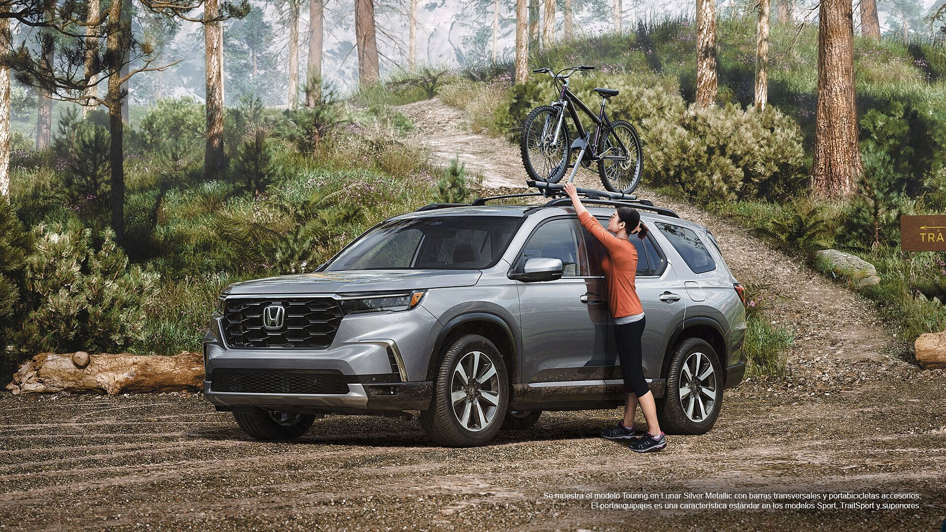 Mujer colocando la bicicleta en el Honda Pilot 2023 mostrado en Lunar Silver metálico con barras transversales accesorias y enganche para bicicleta de pie en un aparcamiento forestal.