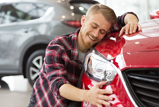 A man wearing a plaid shirt is smiling and hugging the hood of a red car in a showroom.