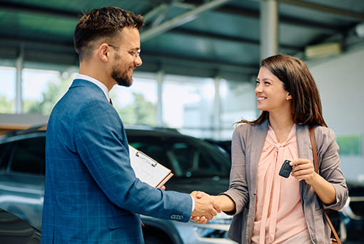 A man in a blue suit and glasses shakes hands with a smiling woman holding car keys in a dealership. They stand by a parked car, conveying satisfaction and trust.