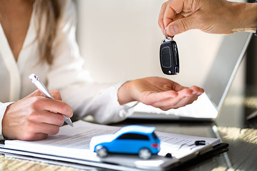 Person handing over car keys to another, who signs a contract on a clipboard. A small blue toy car is on the table, conveying a car sale.