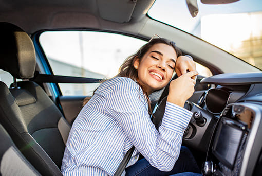 A woman in a striped shirt, seated in a car, joyfully hugs the steering wheel. She smiles with eyes closed, conveying happiness and contentment.