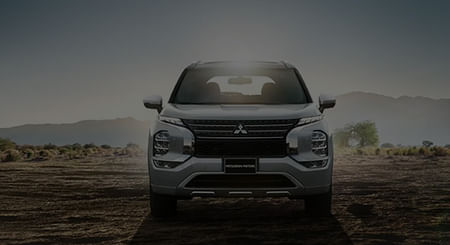 A silver 2025 Mitsubishi Outlander PHEV park in front of a mountainous landscape with the sunset illuminating it.