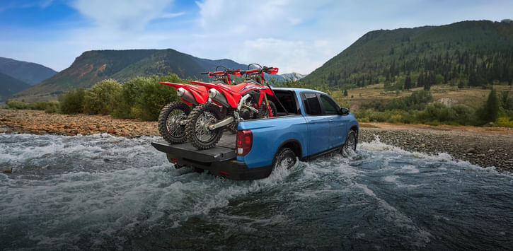 A blue 2025 Ridgeline TrailSport driving across a small river with 2 motorbikes in the tuck bed.