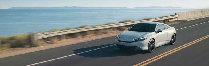 A sleek, silver 2026 Honda Prelude coupe is shown in motion on a sunlit coastal highway, with a vast blue ocean and clear sky in the background.