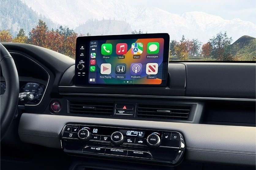 Interior del Honda Pilot 2025 con pantalla táctil digital que muestra iconos de aplicaciones para música, navegación y mensajería, con un fondo escénico.