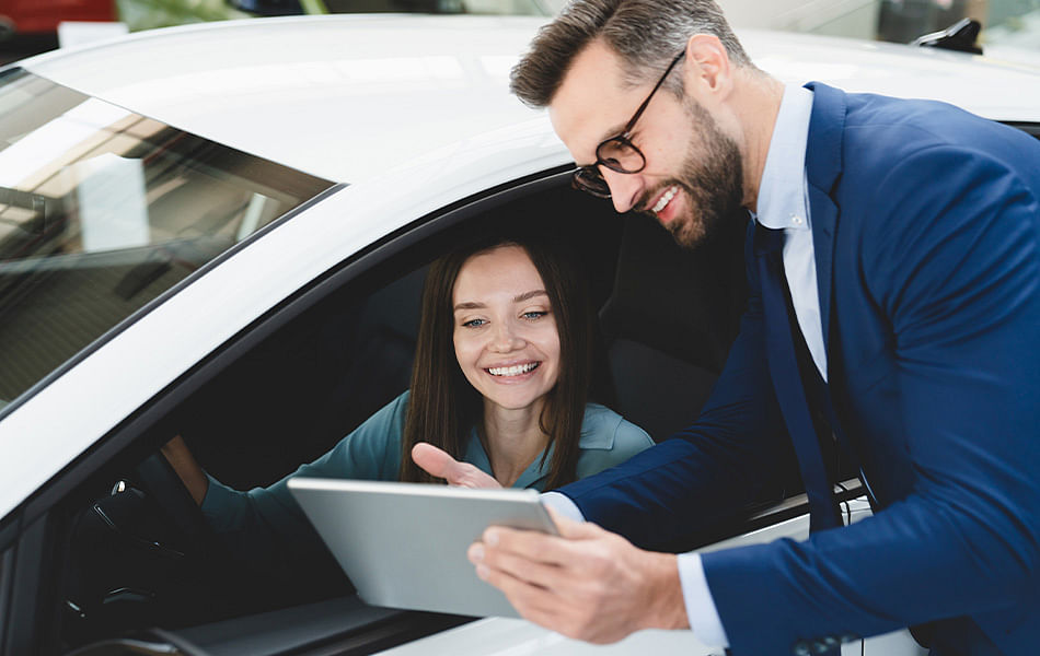 A car dealer stands outside a vehicle while a customer inside examines a tablet, discussing options together.