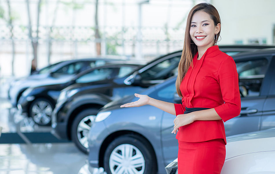 Asian woman in a red dress stands beside luxury cars in a modern showroom, showcasing elegance and style.